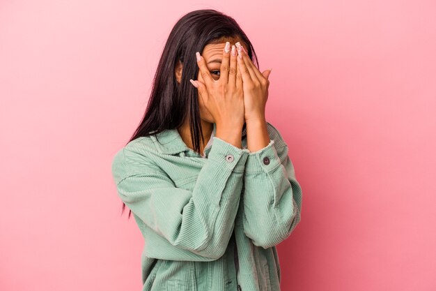 Junge lateinische Frau, die auf rosafarbenem Hintergrund isoliert ist, blinzelt erschrocken und nervös durch die Finger.