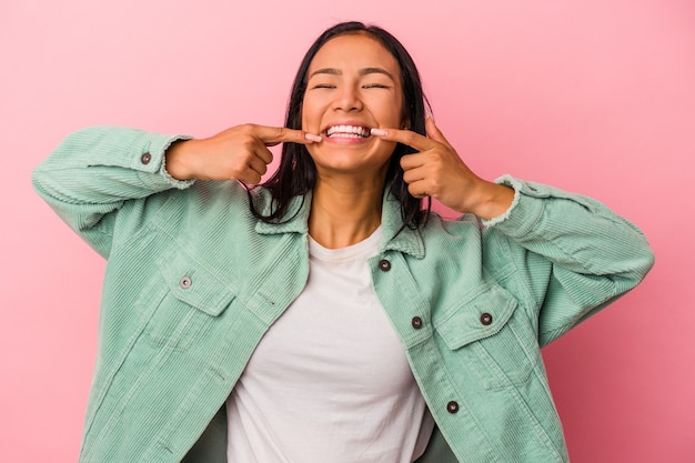 Junge lateinische Frau, die auf rosafarbenem Hintergrund isoliert ist, lächelt und zeigt mit den Fingern auf den Mund.