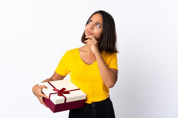 Junge lateinische Frau, die ein Geschenk weiß hält, das Zweifel beim Aufblicken hat