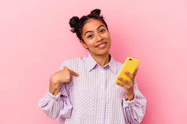 Junge lateinische Frau, die ein Handy lokalisiert auf rosa Hintergrundperson hält, die von Hand auf einen Hemdkopierraum zeigt, stolz und zuversichtlich