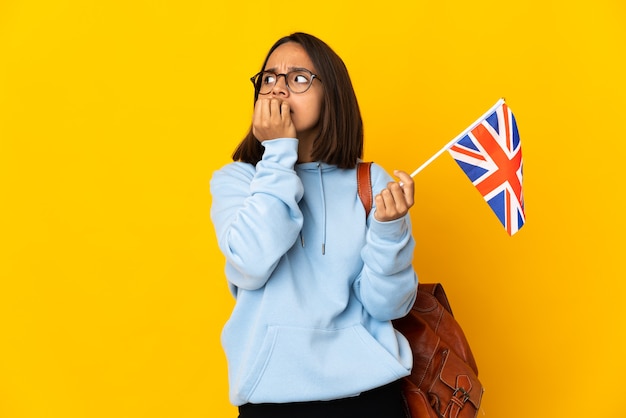 Junge lateinische Frau, die eine britische Flagge lokalisiert auf gelbe Wand nervös und ängstlich hält Hände zu Mund hält