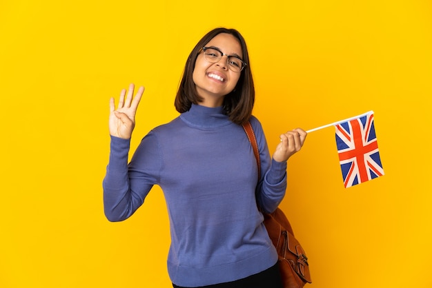 Junge lateinische Frau, die eine britische Flagge lokalisiert auf gelbem Hintergrund glücklich hält und vier mit den Fingern zählt