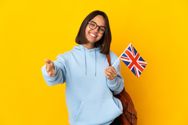 Junge lateinische Frau, die eine britische Flagge lokalisiert auf gelber Wand hält, die präsentiert und einlädt, mit Hand zu kommen