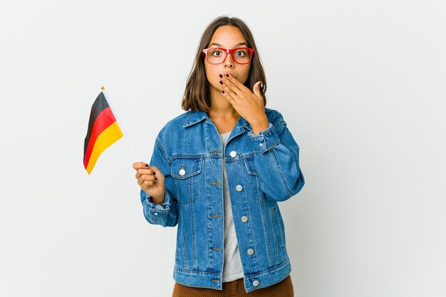 Foto junge lateinische frau, die eine deutsche flagge lokalisiert auf weißem hintergrund hält, schockiert, den mund mit den händen zu bedecken.