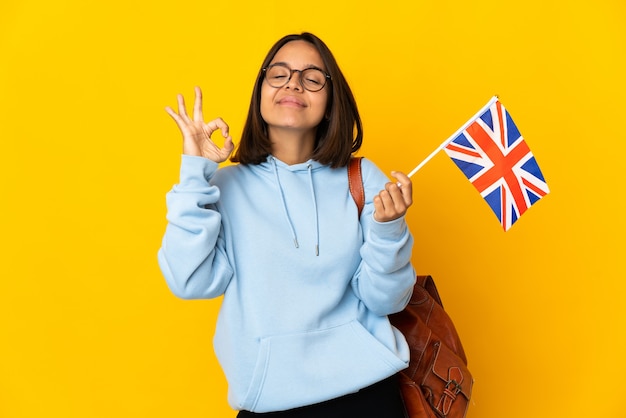 Junge lateinische Frau, die eine Flagge des Vereinigten Königreichs lokalisiert auf gelbem Hintergrund in Zen-Pose hält