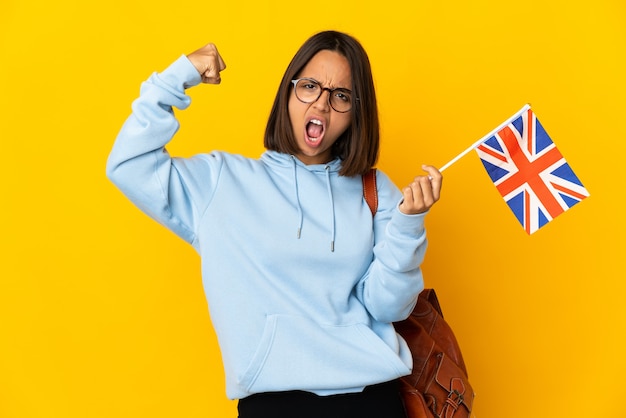 Junge lateinische Frau, die eine Flagge des Vereinigten Königreichs lokalisiert auf gelber Wand hält, die starke Geste tut