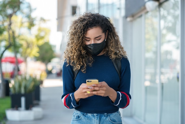 Junge lateinische frau, die eine gesichtsmaske trägt, während sie ihr handy draußen auf der straße benutzt. Neues normales Lifestyle-Konzept. Urbanes Konzept.