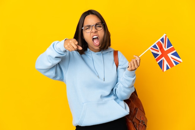 Junge lateinische Frau, die eine Großbritannienflagge lokalisiert auf gelbem Hintergrund frustriert hält und auf die Front zeigt