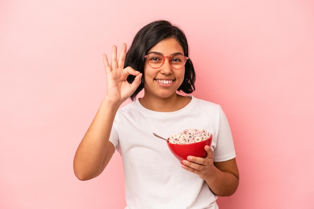 Junge lateinische Frau, die Getreide einzeln auf rosafarbenem Hintergrund hält, fröhlich und selbstbewusst, die eine ok Geste zeigen.