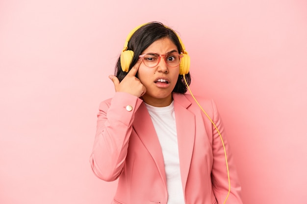 Junge lateinische Frau, die Musik hört, die auf rosafarbenem Hintergrund isoliert ist und eine Enttäuschungsgeste mit dem Zeigefinger zeigt.