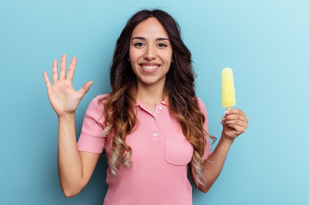Junge lateinische Frau mit Eis auf blauem Hintergrund isoliert lächelnd fröhlich mit Nummer fünf mit den Fingern.