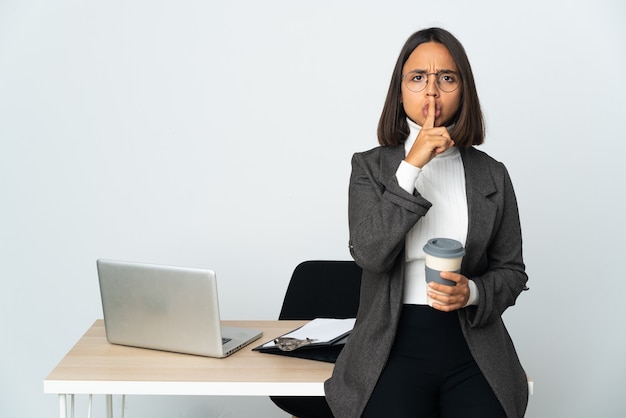 Junge lateinische Geschäftsfrau, die in einem Büro arbeitet, das auf weißem Hintergrund lokalisiert wird, der ein Zeichen der Stille Geste zeigt, die Finger in den Mund setzt