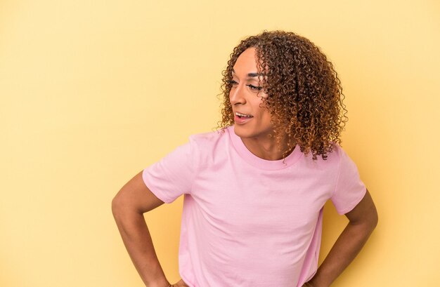 Junge lateinische transsexuelle Frau, die auf gelbem Hintergrund isoliert ist, lacht glücklich und hat Spaß, die Hände auf dem Bauch zu halten.