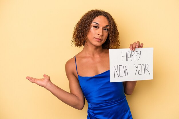 Junge lateinische transsexuelle Frau, die das neue Jahr einzeln auf gelbem Hintergrund feiert, das einen Kopienraum auf einer Handfläche zeigt und eine andere Hand an der Taille hält.