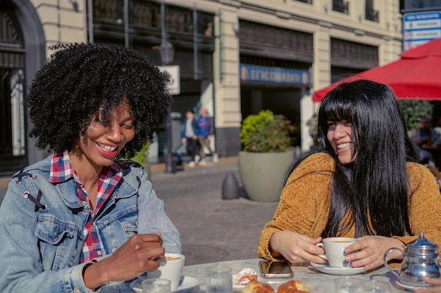 Junge Latina und haitianische Afro-Frau lachen zusammen in einem Café im Freien im Herbst mit einem heißen Getränk