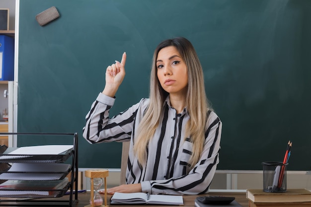 junge lehrerin, die an der schulbank vor der tafel im klassenzimmer sitzt und die hausaufgaben der schüler überprüft, die den zeigefinger zeigen und zuversichtlich aussehen, eine neue idee zu haben