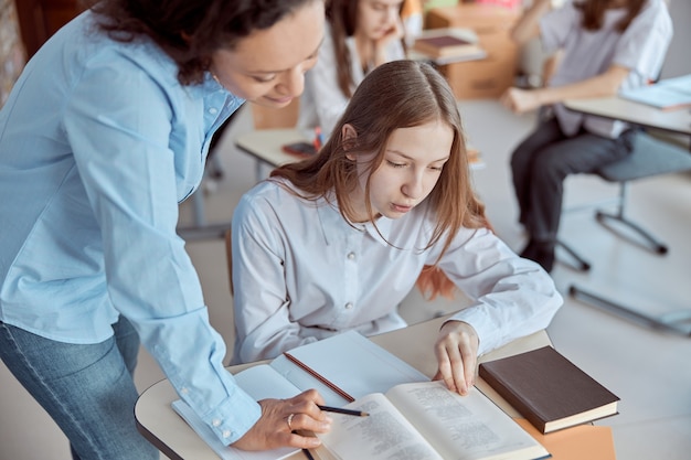 Foto junge lehrerin hilft, ihre schülerin zu lesen. grundschulkinder sitzen auf schreibtischen und lesen bücher im klassenzimmer.