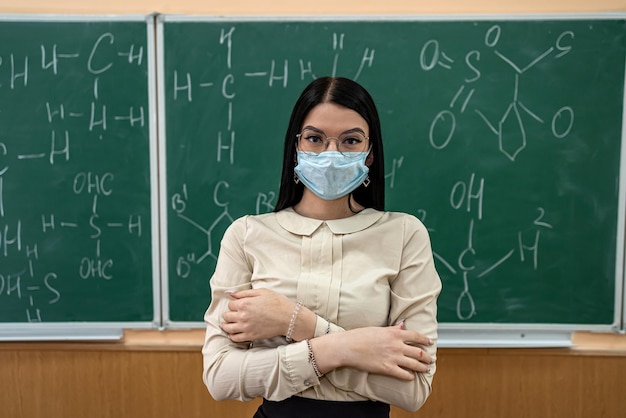 Junge Lehrerin in Schutzmaske, die neben der Tafel steht