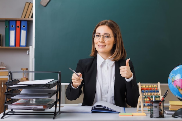 Junge Lehrerin mit Brille, die an der Schulbank vor der Tafel im Klassenzimmer sitzt, mit Abakus und Globus, der das Klassenbuch überprüft und den Zeiger hält, der den Daumen lächelnd nach oben zeigt