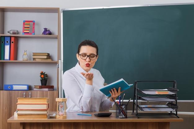 Junge Lehrerin mit Brille, die an der Schulbank vor der Tafel im Klassenzimmer sitzt und ein Buch hält, das glücklich und positiv einen Luftkuss sendet