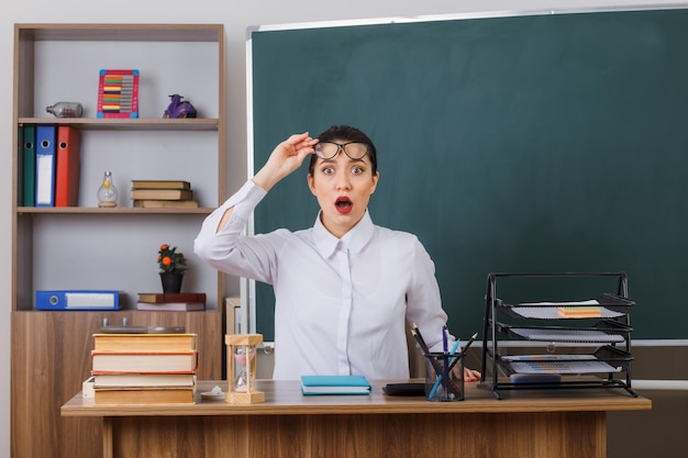 Junge Lehrerin mit Brille, die den Unterricht erklärt und in die Kamera blickt, sitzt erstaunt und schockiert an der Schulbank vor der Tafel im Klassenzimmer