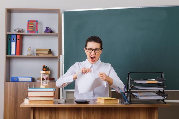 Junge Lehrerin mit Brille, die ein Stück Papier zerreißt und mit aggressivem Ausdruck schreit, sitzt an der Schulbank vor der Tafel im Klassenzimmer