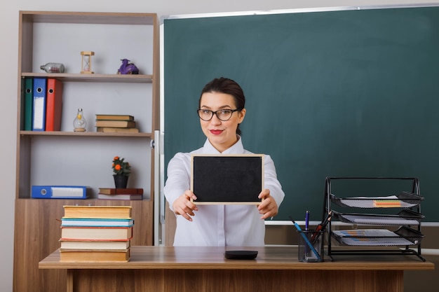 Junge Lehrerin mit Brille, die eine kleine Tafel zeigt, die selbstbewusst lächelt und an der Schulbank vor der Tafel im Klassenzimmer sitzt