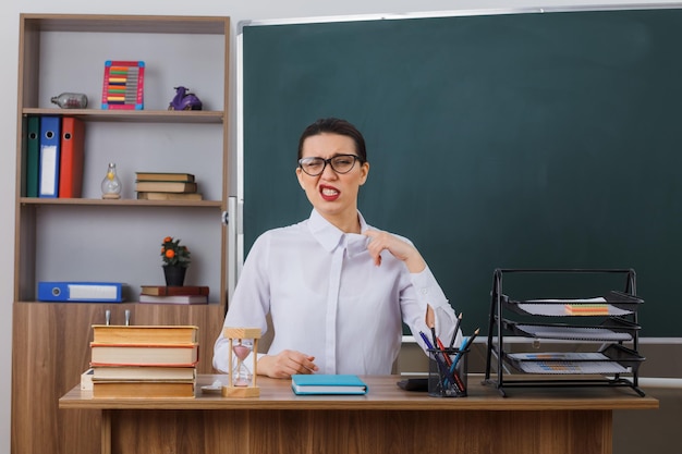 Junge Lehrerin mit Brille, die genervt und irritiert an der Schulbank vor der Tafel im Klassenzimmer sitzt