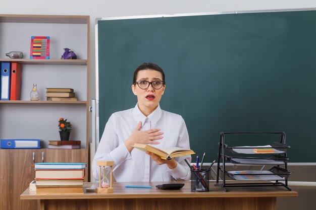 Junge Lehrerin mit Brille sitzt an der Schulbank mit Buch vor der Tafel im Klassenzimmer und blickt besorgt und verwirrt in die Kamera