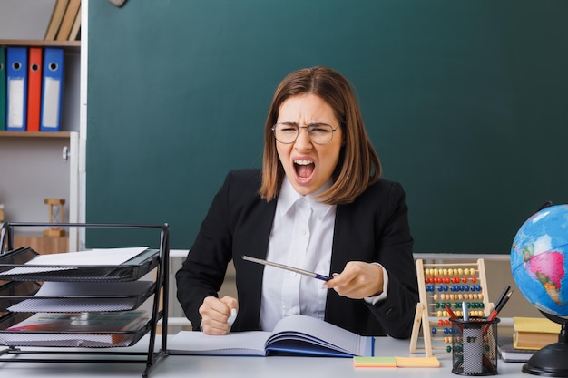 Junge Lehrerin mit Brille sitzt an der Schulbank vor der Tafel im Klassenzimmer mit Abakus, der das Klassenregister überprüft und den Zeiger wütend und wütend mit aggressivem Ausdruck schreit