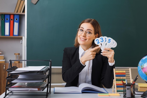 Junge Lehrerin mit Brille sitzt an der Schulbank vor der Tafel im Klassenzimmer und hält Nummernschilder, die den Unterricht glücklich und zufrieden lächelnd erklären