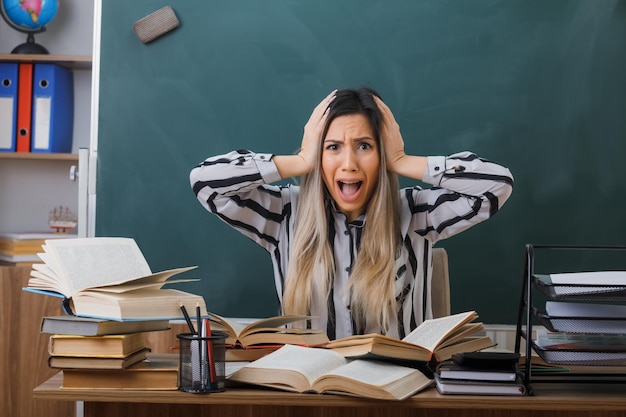 junge lehrerin sitzt an der schulbank vor der tafel im klassenzimmer mit büchern, die auf das chaos auf ihrem schreibtisch blicken und verrückt wütend schreien in panik, die hände auf dem kopf halten