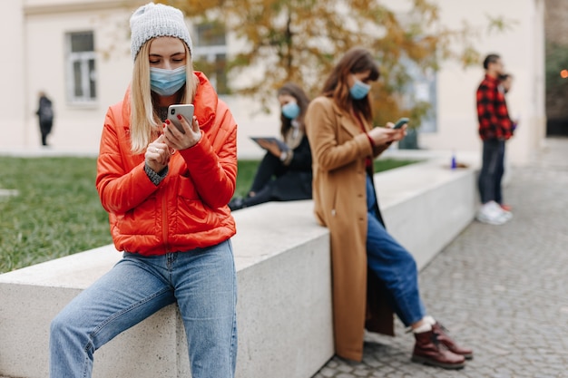 Junge Leute, die moderne Geräte auf der Straße benutzen, medizinische Schutzmasken tragen und Abstand halten. Konzept der Pandemiezeit.