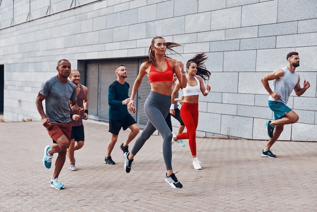 Junge Leute in Sportkleidung joggen in voller Länge, während sie im Freien auf dem Bürgersteig trainieren