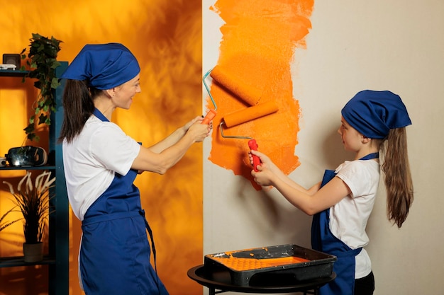 Junge Leute streichen zu Hause orangefarbene Wände und arbeiten mit Farbfarbe und Walzenpinsel an der Wohnungsrenovierung. Frau mit kleinem Kind, die den Hausinnenraum mit Werkzeugen umdekoriert.