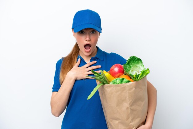 Junge Lieferfrau, die eine Tüte mit Essen zum Mitnehmen isoliert auf weißem Hintergrund nimmt, überrascht und schockiert, während sie nach rechts schaut