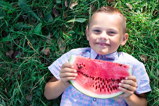 Junge liegt auf grünem Gras und hält ein großes Stück Wassermelone, ein kleiner Schüler isst eine Wassermelone an einem sonnigen Sommertag, Kopienraum