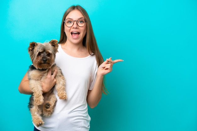 Junge litauische Frau, die einen Hund isoliert auf blauem Hintergrund hält, überrascht und zeigt mit dem Finger zur Seite