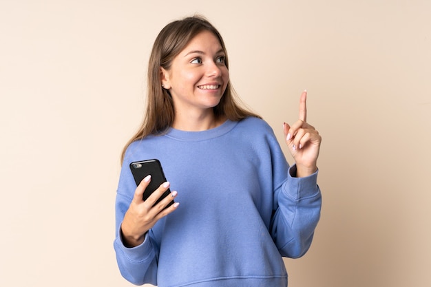 Junge litauische Frau, die Handy verwendet, das auf beige Wand lokalisiert ist, die beabsichtigt, die Lösung zu realisieren, während sie einen Finger anhebt