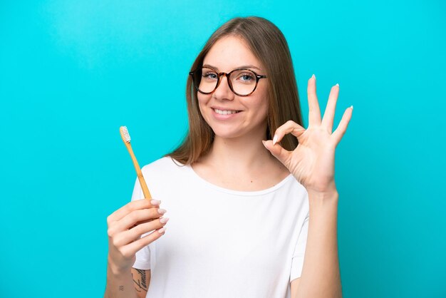 Junge litauische Frau putzt sich die Zähne vor isoliertem Hintergrund und zeigt mit den Fingern ein Ok-Zeichen