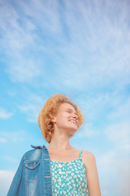 junge lockige rothaarige frau in strohhut blauem sommerkleid und jeansjacke, die auf blauem himmel steht