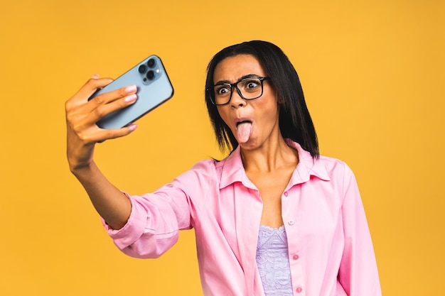 Junge, lustige, afrikanisch-amerikanische, schwarze, lässige Frau, die das Gesicht verzieht und Selfie auf dem Smartphone macht, isoliert über gelbem Hintergrund