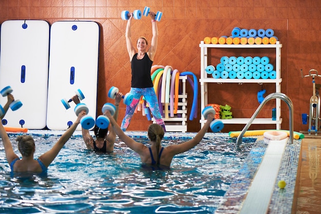 Junge Mädchen und Frauen mittleren Alters machen Aqua-Aerobic mit Hanteln in einem Schwimmbad mit einem Trainer Fitness-Übungen zur Gewichtsreduktion im Wasser