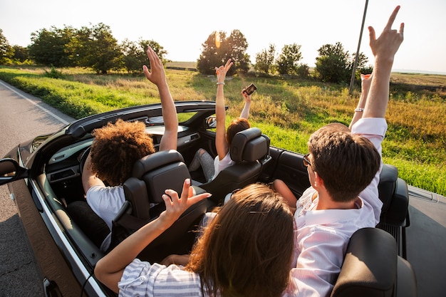 Junge Mädchen und Jungs sitzen in einem schwarzen Cabriolet, halten ihre Hände hoch und machen an einem warmen sonnigen Tag Selfies. .