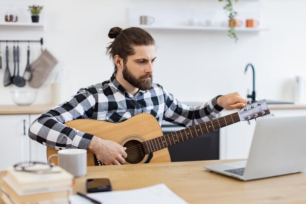 Junge männliche Musiker stimmen Gitarre mit einem Laptop auf einem Schreibtisch in einer modernen Wohnung