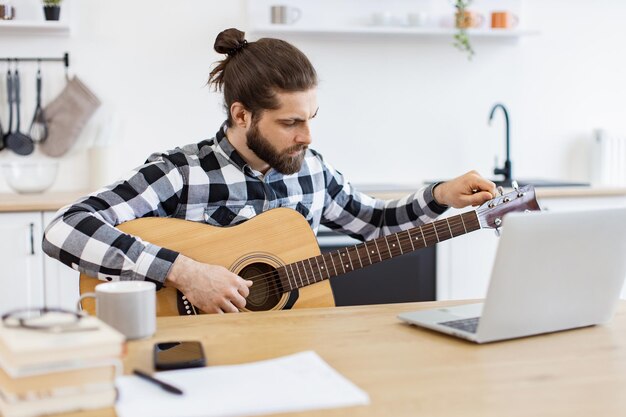 Junge männliche Musiker stimmen Gitarre mit einem Laptop auf einem Schreibtisch in einer modernen Wohnung