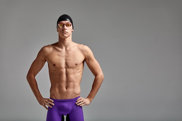 Junge männliche Schwimmer, die sich auf den Start vorbereiten, halblanges Porträt eines Schwimmers in Maske und Hut, grauer Hintergrund, Kopierraum, Schwimmkonzept.