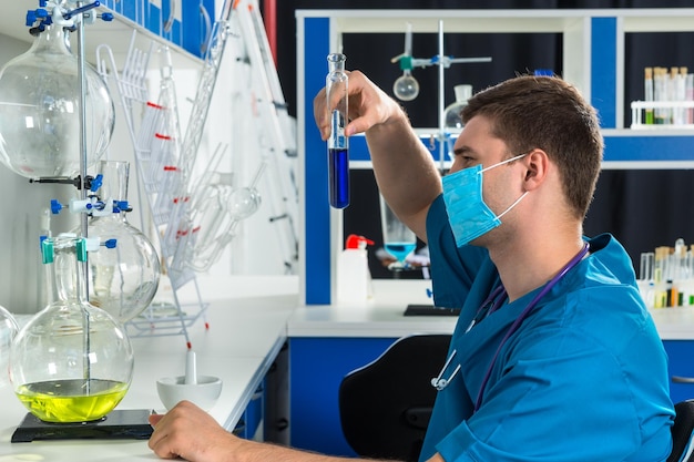 Junge männliche Wissenschaftler in Uniform, die eine Maske tragen, halten ein großes Reagenzglas mit blauer Flüssigkeit und forschen in einem Labor. Gesundheits- und Biotechnologiekonzept