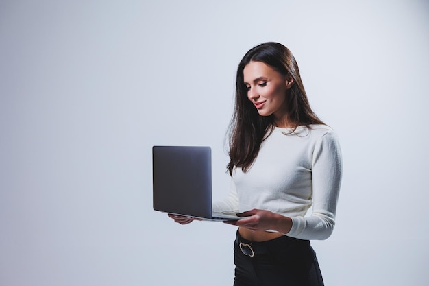 Junge Managerin in einem weißen Hemd hält einen tragbaren Laptop in ihren Händen, während sie auf einem grauen Hintergrund steht