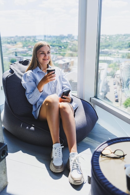 Junge Managerin mit Kopfhörern, die telefoniert und Kaffee trinkt, während sie in einem modernen Coworking Space sitzt Freiberufliches Mädchen, das remote online arbeitet